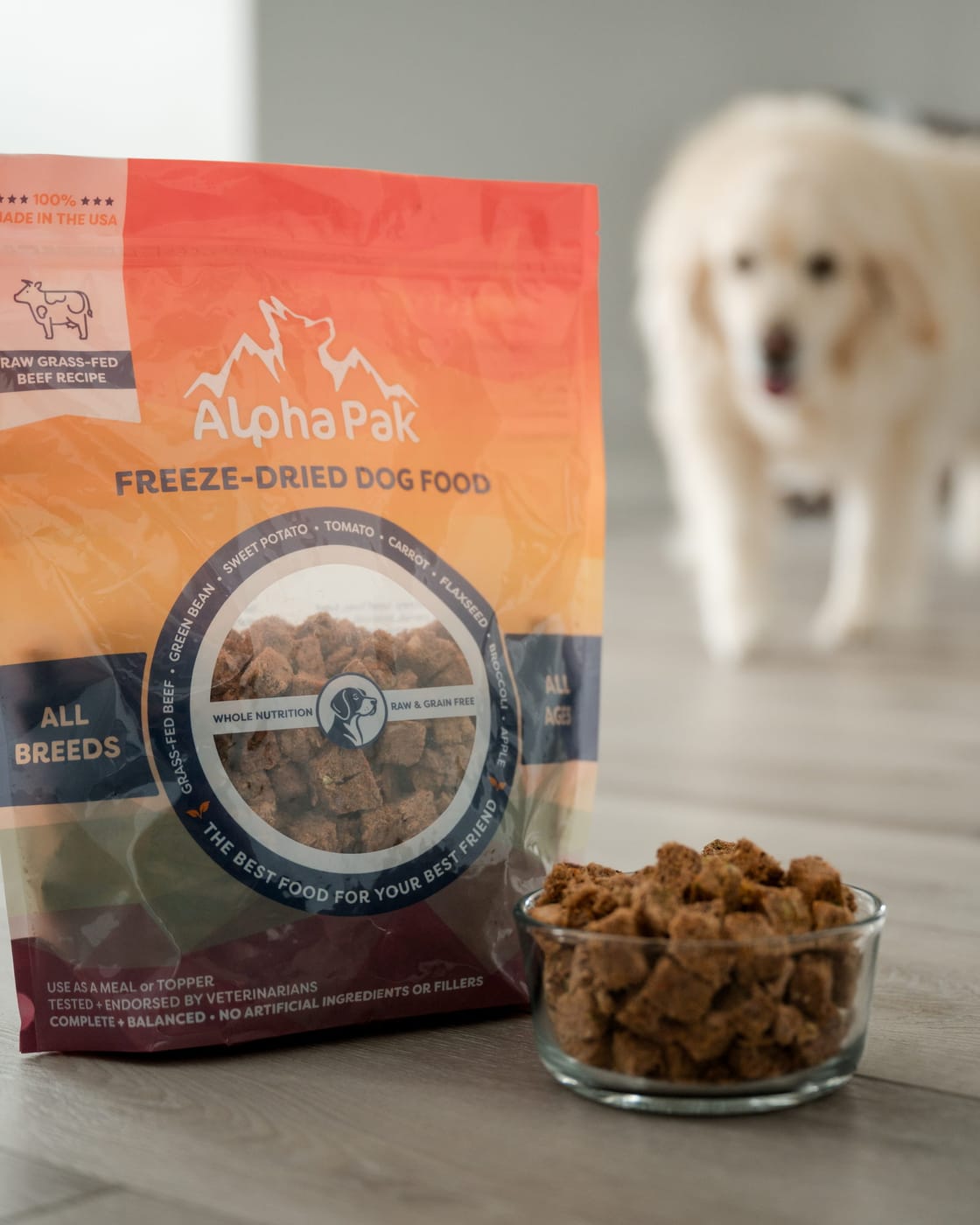 An AlphaPak beef recipe bag beside a bowl of freeze-dried raw food on a wood floor, a cream golden retriever walking toward it